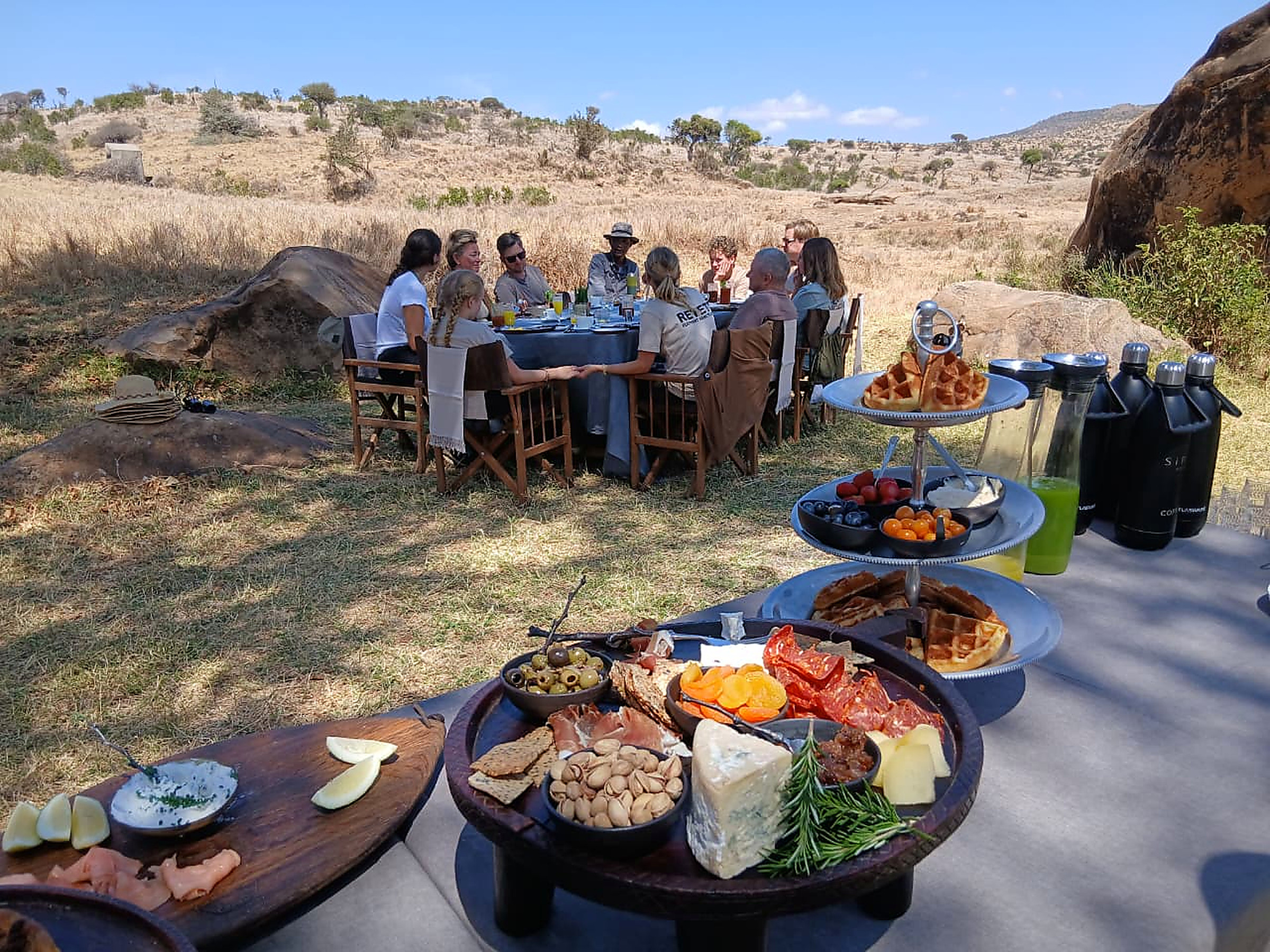 Guests seated at a long table for a bush breakfast in the open, with a spread of food in the foreground and dry hills behind