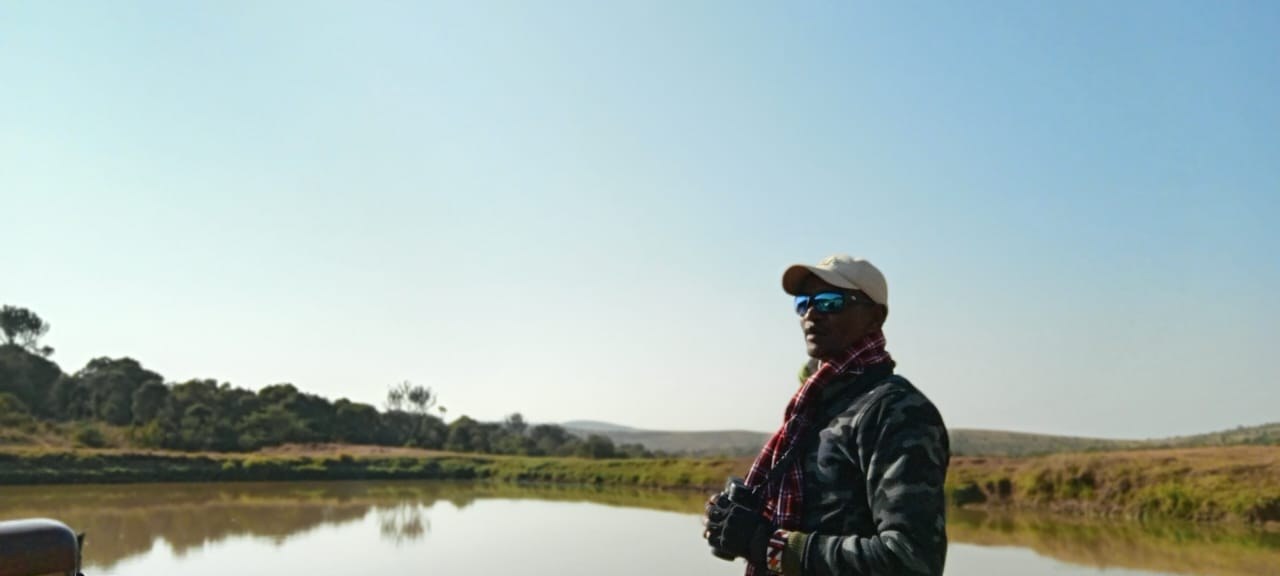 A person in a cap and jacket standing beside still water at dawn, binoculars in hand