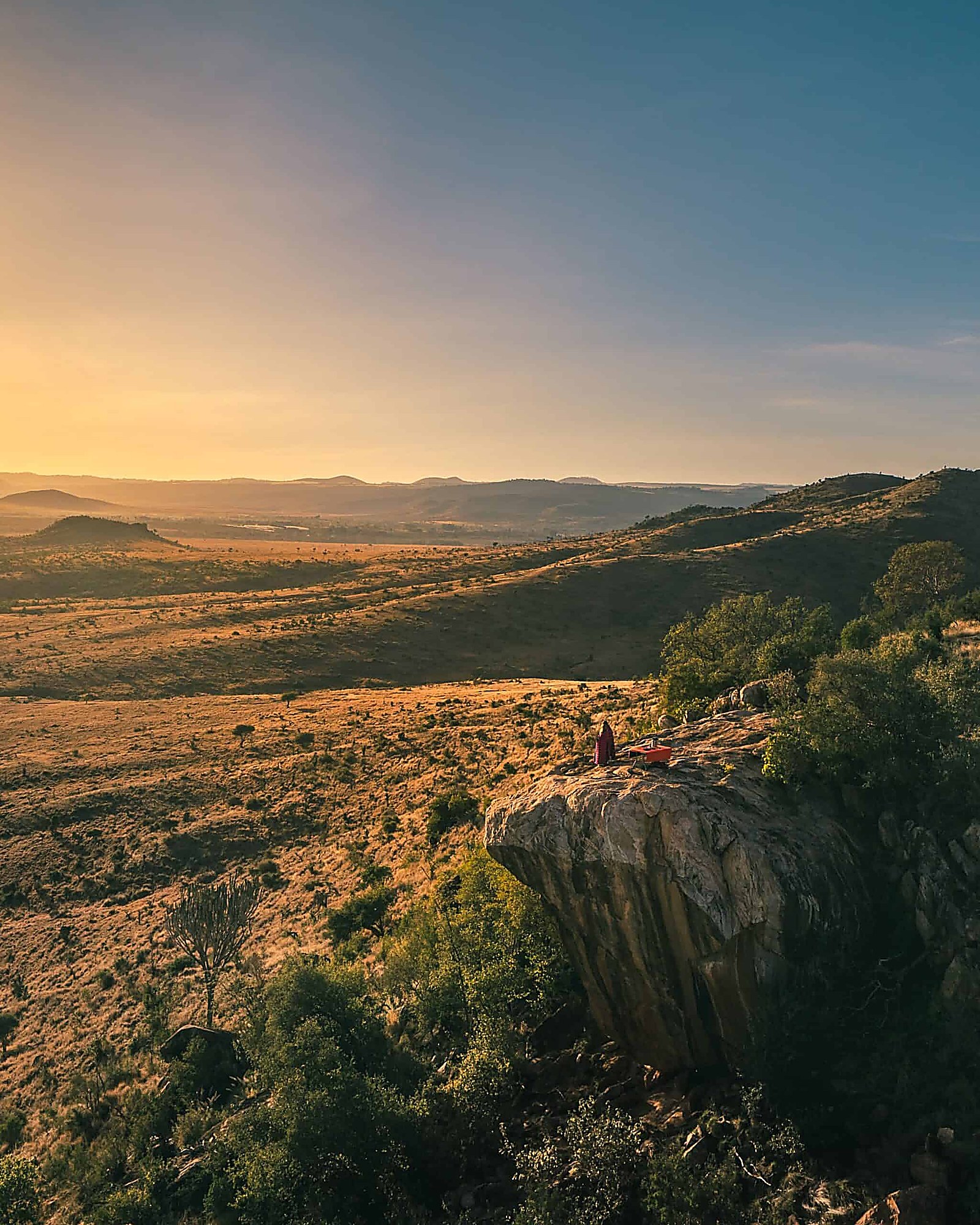 A figure in a red shawl standing on a rock outcrop above a wide valley at dawn