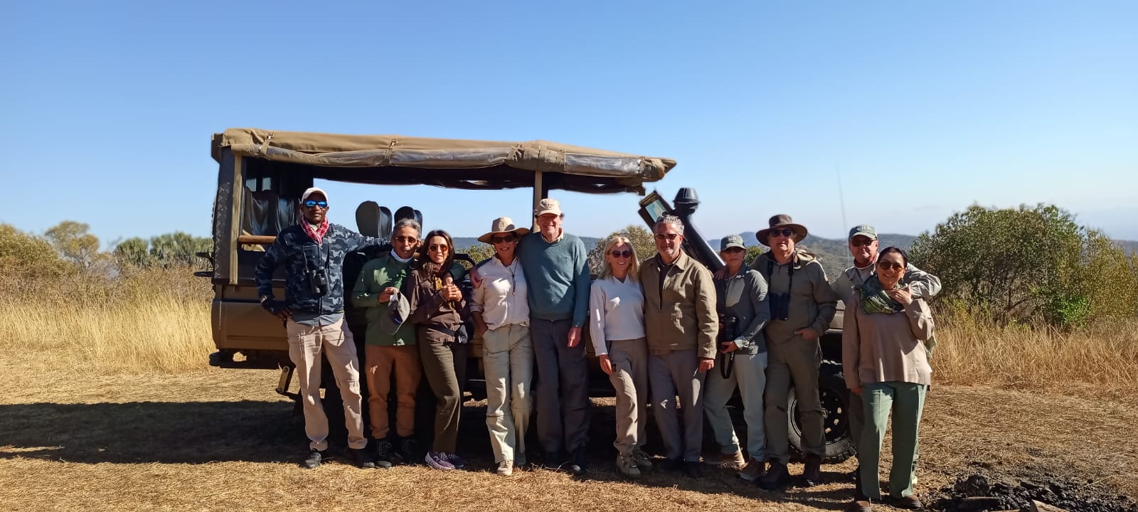 A safari group standing together with their guide in front of an open-sided safari vehicle, dry golden grass and open bush behind them